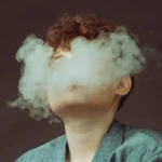 "Person exhaling a dense cloud of vapor with face obscured, symbolizing the use of a 'Terp Ready Ball Vape' device. The image highlights the effect of vaporization, showcasing a thick, cloudy exhale against a muted background."