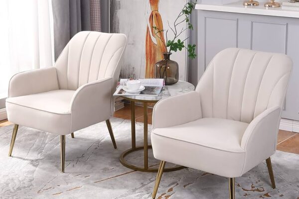 Elegant set of two white accent chairs with gold metal legs, placed around a small round marble table in a stylish living room setting. Decor includes a modern painting, vase with greenery, and candles on a mantel