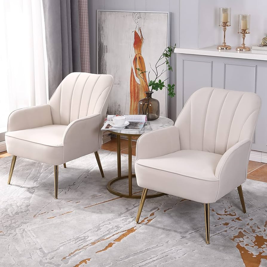 Elegant set of two white accent chairs with gold metal legs, placed around a small round marble table in a stylish living room setting. Decor includes a modern painting, vase with greenery, and candles on a mantel