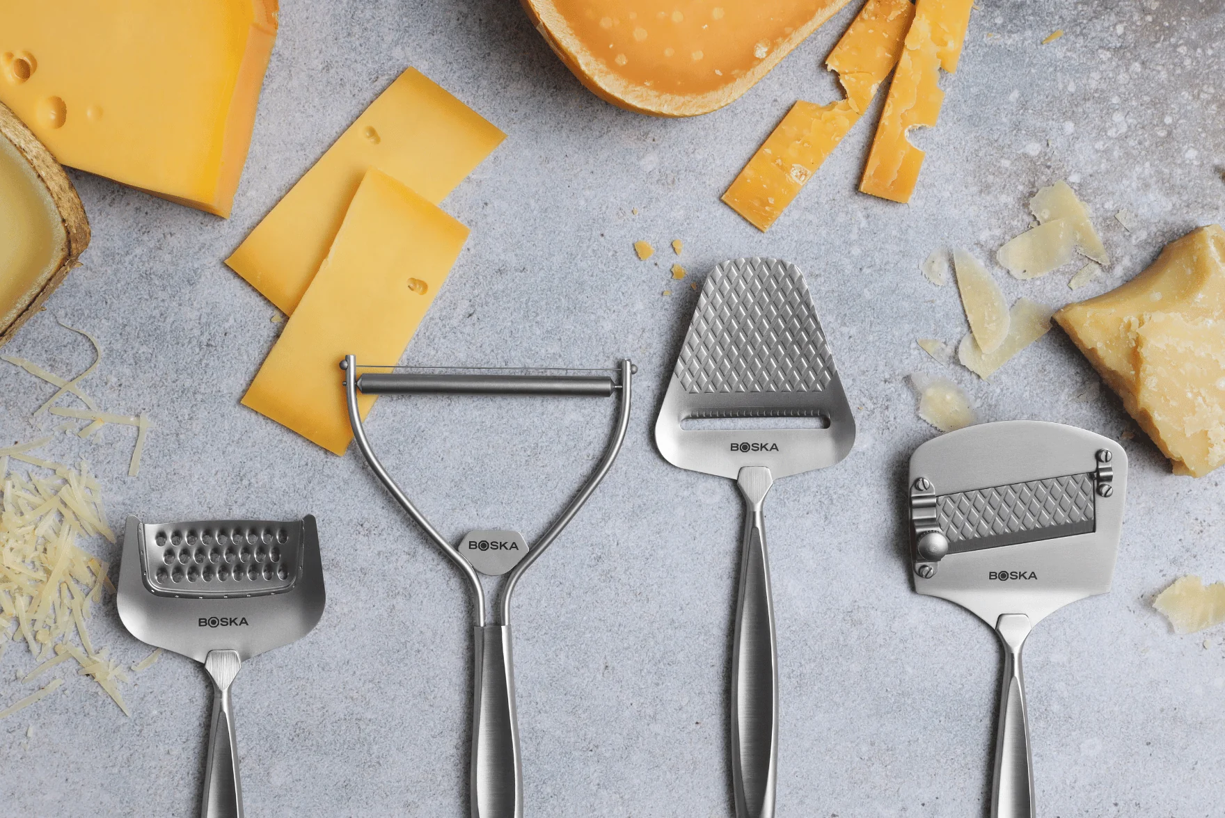 A selection of stainless steel Boska Cheese Slicers tools, including slicers and graters, displayed on a gray surface alongside various types of cheese, such as sliced, shredded, and broken chunks of hard cheese.