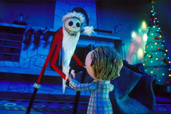 Scene from a stop-motion animated film featuring a skeletal character dressed as Santa Claus interacting with a child in pajamas. The character has a long, red Santa suit, a white beard, and a skeletal face, while the child is holding a gift. A decorated Christmas tree with colorful lights is in the background, adding a festive atmosphere.