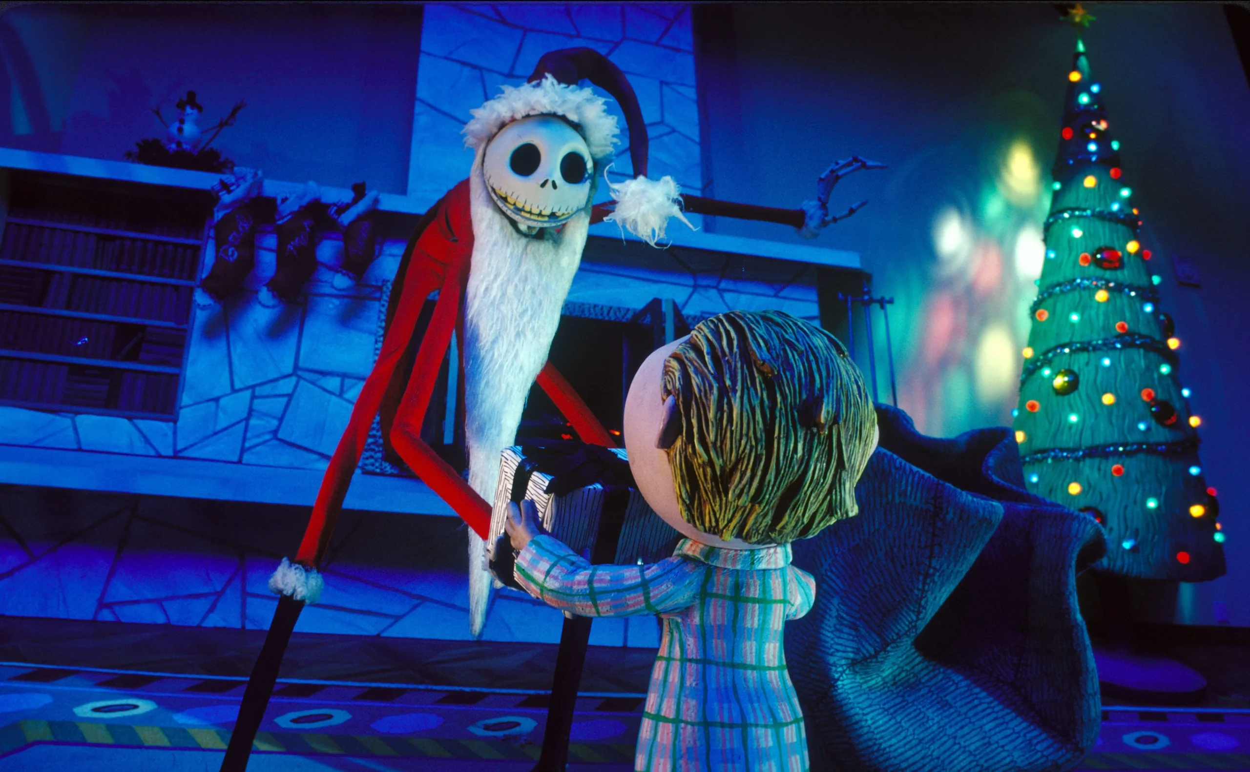 Scene from a stop-motion animated film featuring a skeletal character dressed as Santa Claus interacting with a child in pajamas. The character has a long, red Santa suit, a white beard, and a skeletal face, while the child is holding a gift. A decorated Christmas tree with colorful lights is in the background, adding a festive atmosphere.