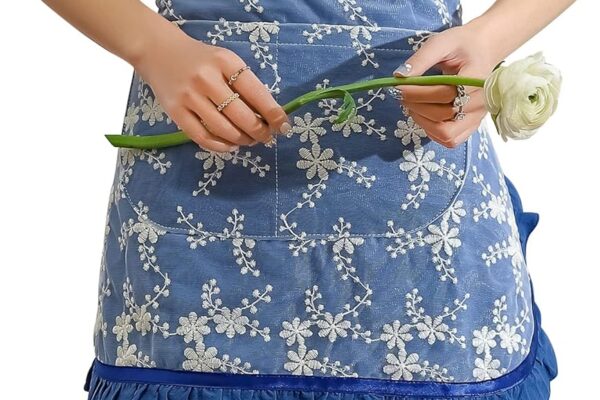 Person wearing a blue Women's Half Aprons with white floral embroidery and ruffled hem, holding a white flower with both hands.