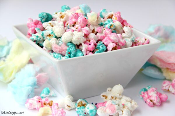 A white bowl filled with colorful cotton candy-flavored popcorn, coated in pastel shades of pink, blue, and white, with additional pieces of popcorn scattered around the bowl on a white surface.