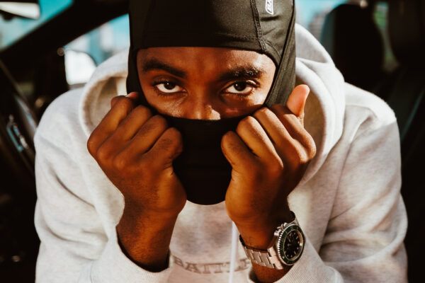 Close-up of a person wearing a black balaclava mask, partially covering their face, with hands held up near the mask. They are dressed in a light gray hoodie and wearing a silver wristwatch, sitting inside a vehicle