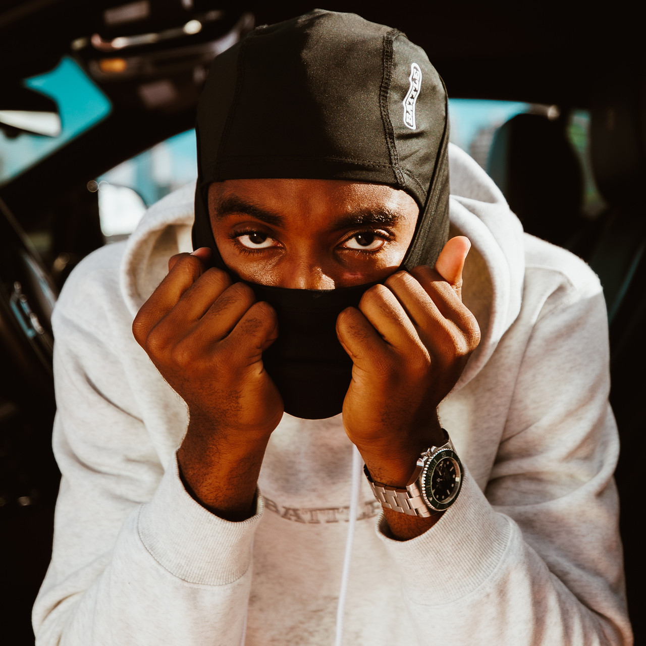 Close-up of a person wearing a black balaclava mask, partially covering their face, with hands held up near the mask. They are dressed in a light gray hoodie and wearing a silver wristwatch, sitting inside a vehicle