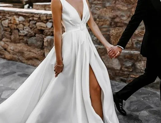 Bride in a flowing white satin dress gown with a deep V-neckline and thigh-high slit, holding hands with a groom in a black suit against a rustic stone wall backdrop