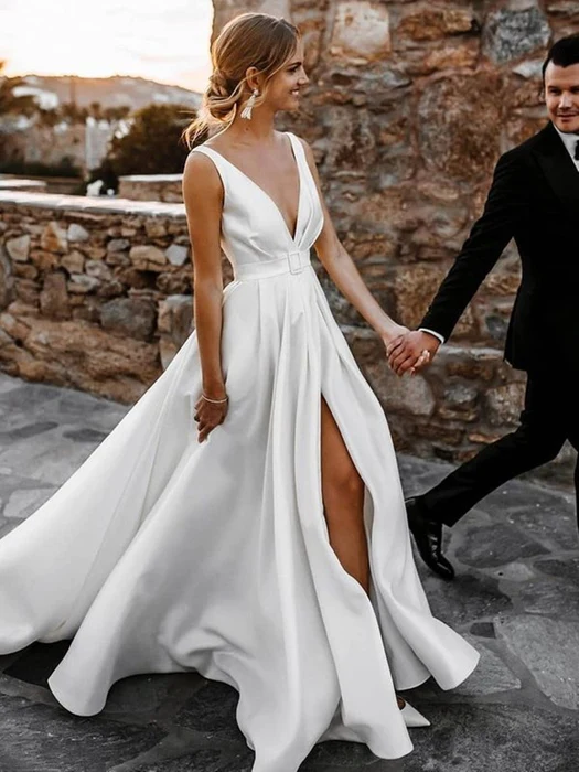 Bride in a flowing white satin dress gown with a deep V-neckline and thigh-high slit, holding hands with a groom in a black suit against a rustic stone wall backdrop