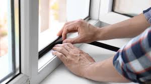 Close-up image of hands applying weatherstripping tape to a window frame for insulation. The person is wearing a plaid shirt, and the scene emphasizes a home improvement task aimed at sealing gaps around the window.