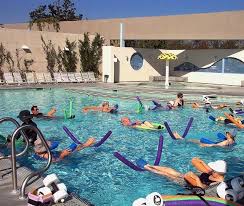 Group of people participating in a Simpkins Swim Center aerobics class at an outdoor pool, using colorful pool noodles for support.