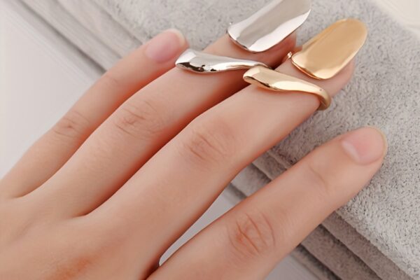 Close-up image of hands adorned with gold fingernail rings on two fingers, showcasing a unique and stylish nail accessory. The hands are neatly manicured, and one wrist is wearing a silver bangle bracelet.