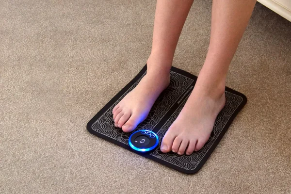 A person with bare feet using an EMS foot massager pad on a carpeted floor. The black pad features a decorative pattern and a circular control panel with a blue LED light, indicating it is powered on.