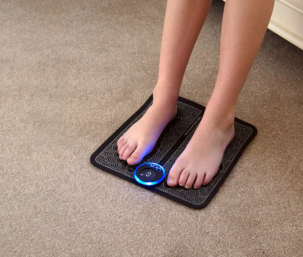 A person with bare feet using an EMS foot massager pad on a carpeted floor. The black pad features a decorative pattern and a circular control panel with a blue LED light, indicating it is powered on.