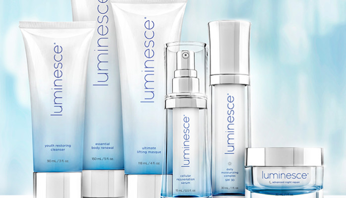 A collection of Luminesce skincare products, including tubes, bottles, and a jar, displayed against a soft blue gradient background. The products are labeled with names such as 'Youth Restoring Cleanser,' 'Essential Body Renewal,' and 'Cellular Rejuvenation Serum.