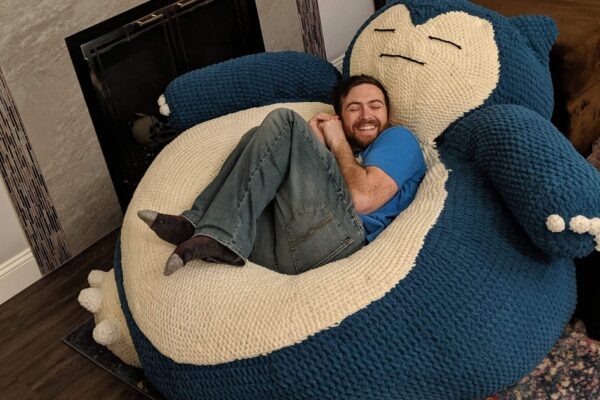 A man with a beard, wearing a blue t-shirt and jeans, is lying comfortably on a large, plush Snorlax bean bag. He is smiling with his eyes closed, appearing relaxed. The Snorlax bean bag is oversized, crocheted, and designed to resemble the Pokémon character Snorlax, with a blue and cream-colored body, arms, and feet. The setting is a cozy living room with a fireplace in the background.