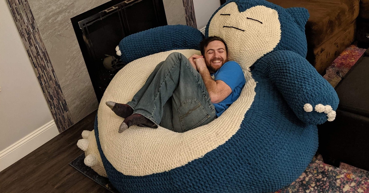 A man with a beard, wearing a blue t-shirt and jeans, is lying comfortably on a large, plush Snorlax bean bag. He is smiling with his eyes closed, appearing relaxed. The Snorlax bean bag is oversized, crocheted, and designed to resemble the Pokémon character Snorlax, with a blue and cream-colored body, arms, and feet. The setting is a cozy living room with a fireplace in the background.