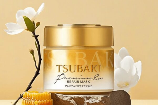 A gold jar of Tsubaki Hair Mask Premium EX Repair Mask displayed on a rustic wooden base, surrounded by white flowers and a honeycomb, set against a warm yellow background.