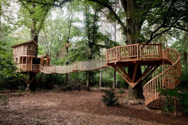 An elaborate wooden Tree Fort Kits fort with multiple levels, a rope bridge, and a spiral staircase, nestled among lush green trees in a forest setting.